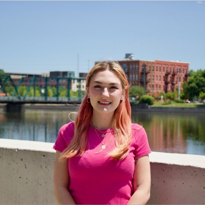 A JMC student with pink hair and a bright pink shirt smiles at the camera while standing near a river. Behind her, there is a green bridge, red brick buildings, trees, and clear blue sky.