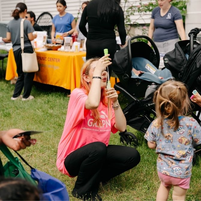 A JMC student in a pink shirt crouches on grass, holding up a green toy while interacting with a young child nearby. Several people, including parents, are gathered around an outdoor table covered with a yellow cloth, suggesting a community event or fair.
