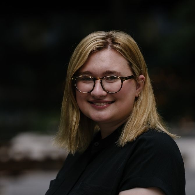 A JMC student with blonde hair and round-framed glasses smiles warmly at the camera. She wears a black shirt, standing against a dark, blurred outdoor background.