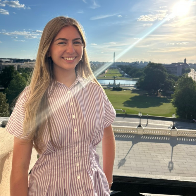 A JMC student with long blonde hair smiles at the camera while standing outdoors. She is wearing a pink and white striped dress, positioned on an elevated terrace overlooking a scenic urban landscape.