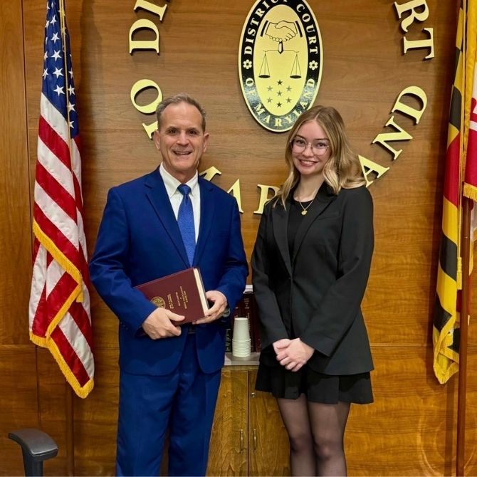 JMC student wearing a black dress and blazer and a judge wearing a navy blue suit, posing together inside of a courthouse.