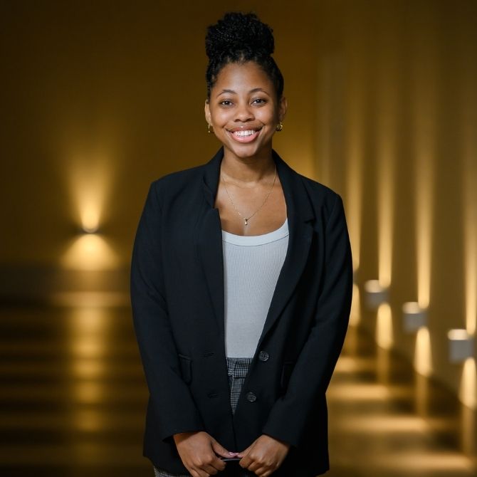 A JMC student smiles warmly while standing confidently in a hallway. She wears a black blazer over a light gray top, accessorized with earrings and a necklace, against a backdrop of warm, golden lighting.