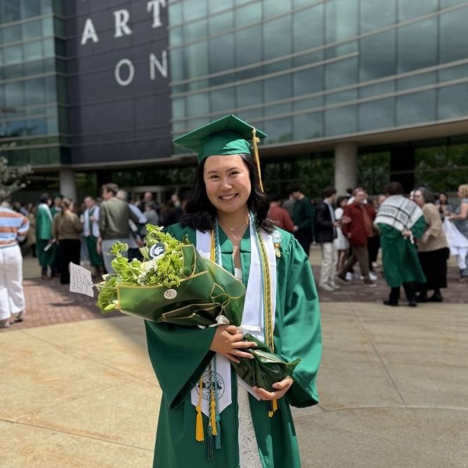 A smiling James Madison College graduate in a green cap and gown holds a bouquet of flowers while standing outside a modern building. Other graduates and attendees are visible in the background.