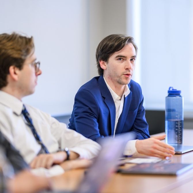 A JMC student wearing a blue suit jacket and white shirt is seated at a conference table, actively speaking with his hands gesturing. Another person sits beside him, partially out of focus, suggesting an ongoing professional discussion or presentation.
