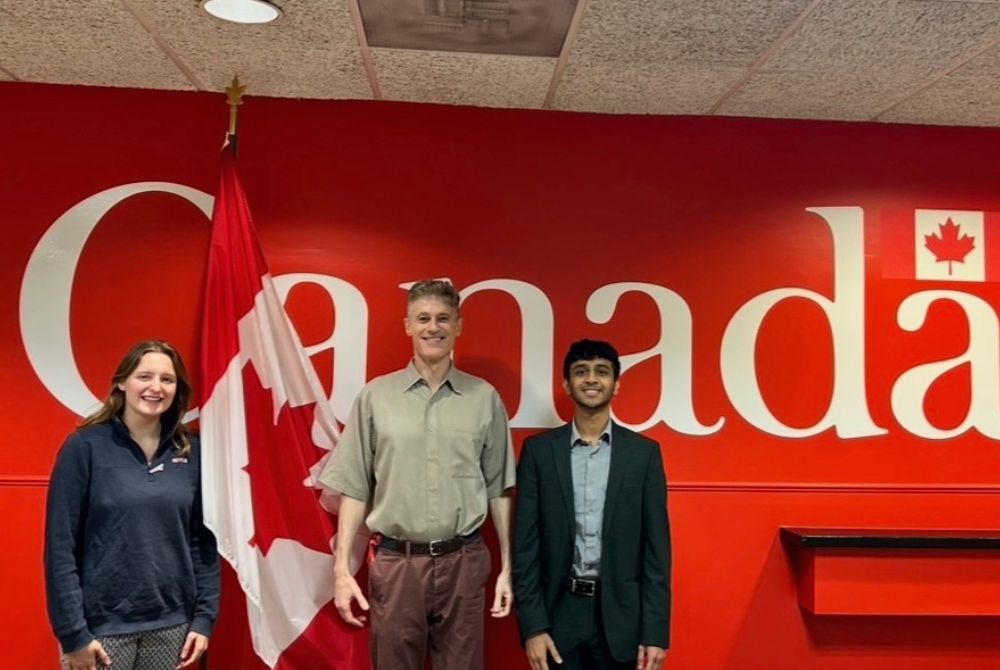 JMC students Grace Fitzgerald and Anish Danappanavar standing with Jan Scazighino, in front of a red wall with Canada spelled out in large white letters.