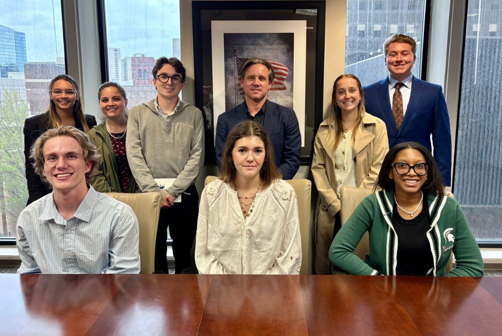 JMC students gathered in a downtown Detroit office conference room, with a framed print of the Declaration of Independence and the city skyline visible behind them.