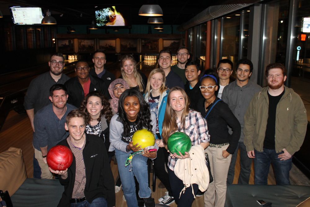 A large group of JMC students poses together at a bowling alley, with three students in front holding red, yellow, and green bowling balls.