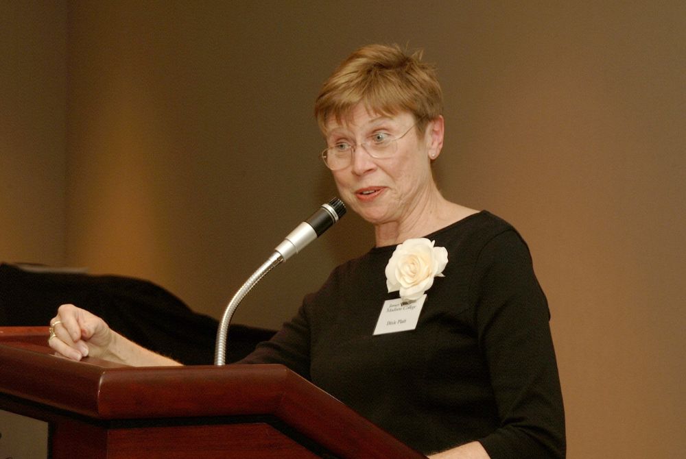 Dixie Platt, with short hair and glasses, is speaking into a microphone while standing behind a wooden podium. She wears a black top, has a white rose pinned to her chest, and appears engaged in delivering a speech or presentation.