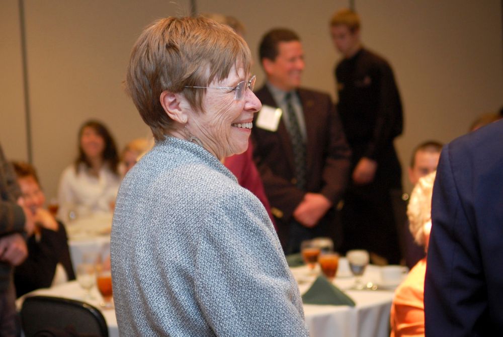 A woman with short hair and glasses smiles while seated at a table during an indoor gathering. Other attendees are visible in the background, some standing, others sitting, suggesting a social or professional event.