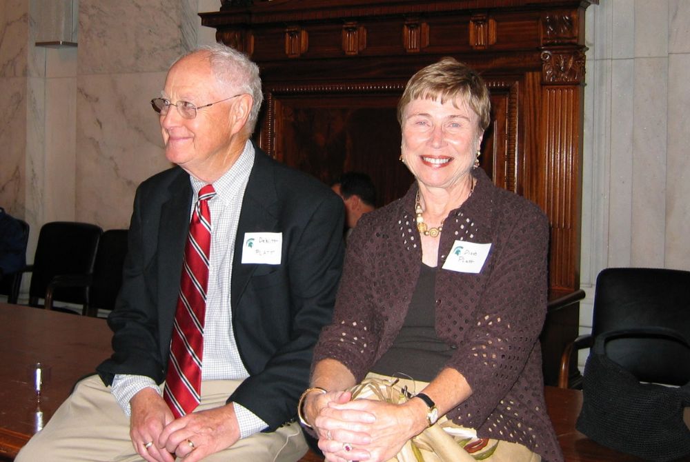 An older man and woman are seated side by side, smiling, in a formal setting with ornate wood paneling. Both wear name tags, suggesting they are attending an organized gathering or conference.