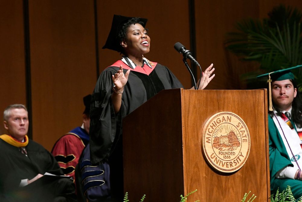 A commencement speaker in academic regalia stands at a podium, speaking into a microphone during what appears to be a graduation ceremony. The wooden podium features the engraved seal of Michigan State University, indicating the event's location.