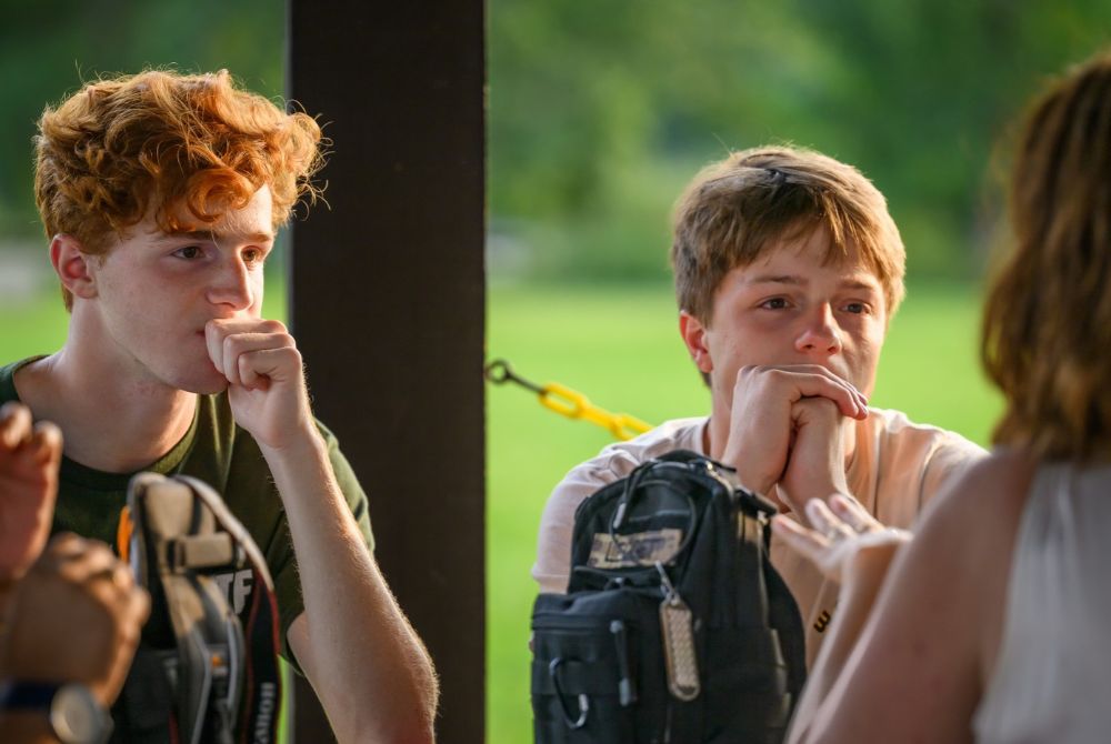 A JMC student with red hair sits thoughtfully, hand near his mouth, while another student looks on, hands clasped together. They are seated outdoors, surrounded by greenery, suggesting a casual, relaxed environment.