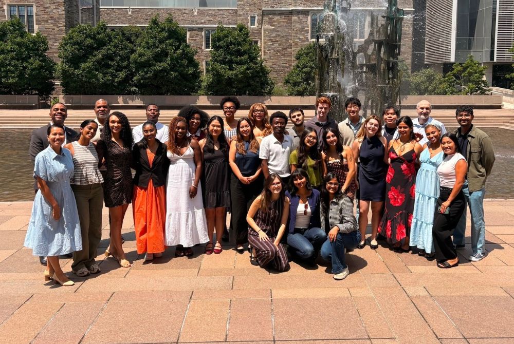 A group of students and faculty pose for a photo in front of a large fountain on a university campus. They are arranged in two rows, with some standing and others kneeling, all smiling toward the camera.