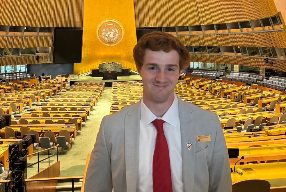 A JMC student in a suit and red tie stands confidently in the United Nations General Assembly hall, smiling slightly. The vast chamber behind him features rows of empty desks, wooden paneling, and the iconic UN emblem on the wall.