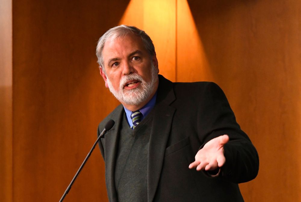 A man with a white beard and gray hair is standing behind a microphone, gesturing as he speaks. He wears a dark suit over a sweater vest, against a warm-toned wooden background.