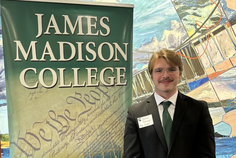 Madison student Parker Dennings, wearing a suit and tie, standing in front of a vertical banner that reads "James Madison College."