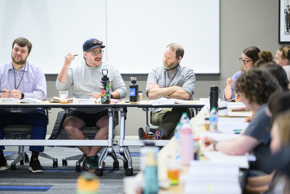 A group of teacher participants in discussion during the JMC Academy for Civic Education. They are seated at tables, with work books and water bottles visible across the desks.