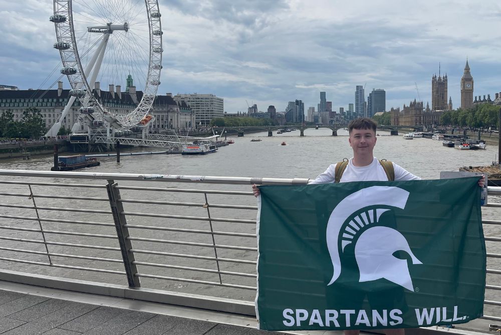 MSU graduate standing on a bridge holding a Spartans Will flag that features the Spartan helmet. In the background is the London Eye ferris wheel.