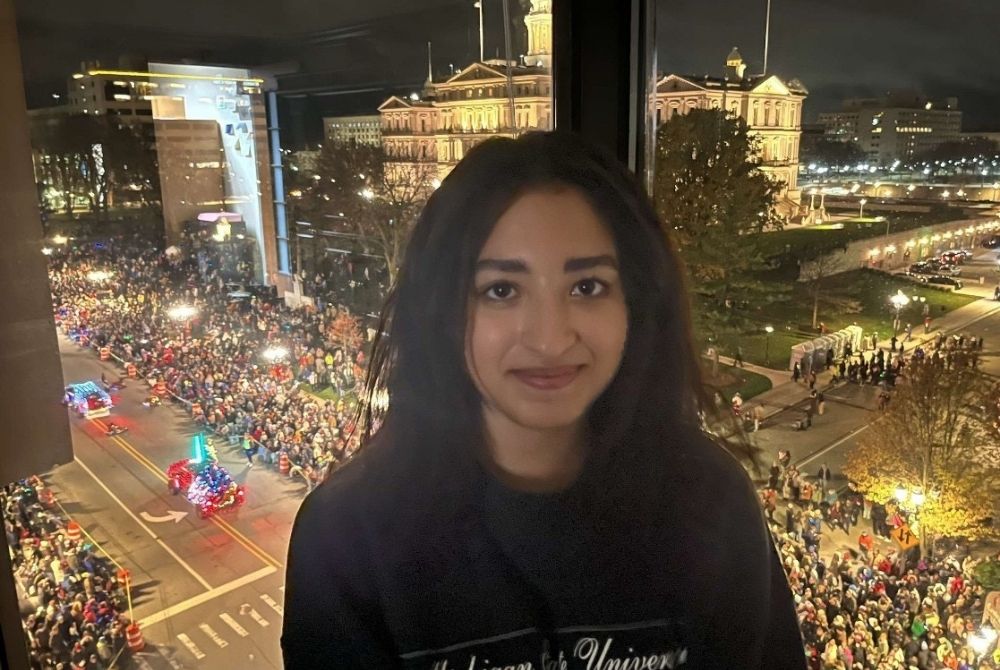 with dark hair smiles at the camera while standing indoors, overlooking a vibrant nighttime parade filled with crowds and colorful floats. The scene outside features illuminated buildings, including a prominent historic structure, under artificial lighting.