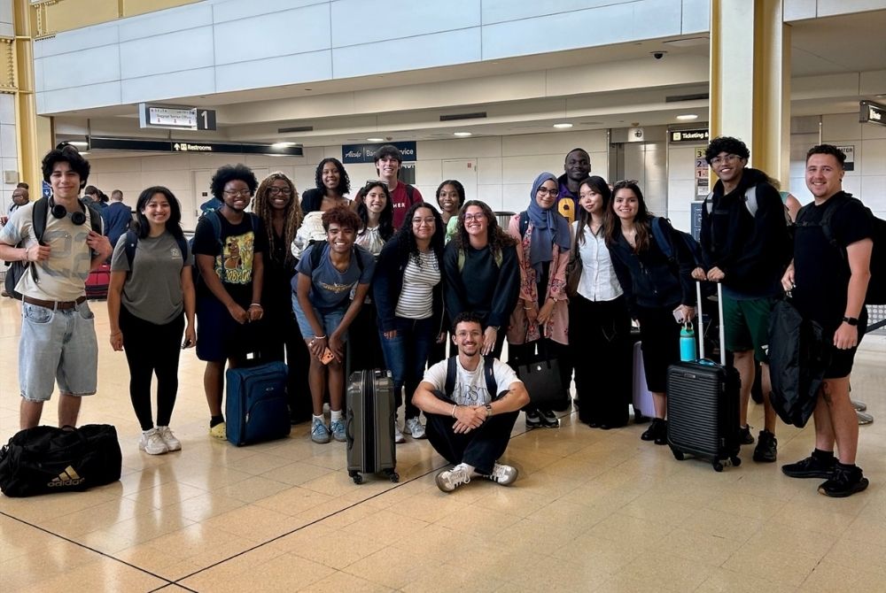 A group of young students, some kneeling and others standing, pose for a photo in an airport terminal while holding or standing next to their luggage. The setting includes visible signs, tiled floors, and overhead lighting, indicating they are preparing for travel.