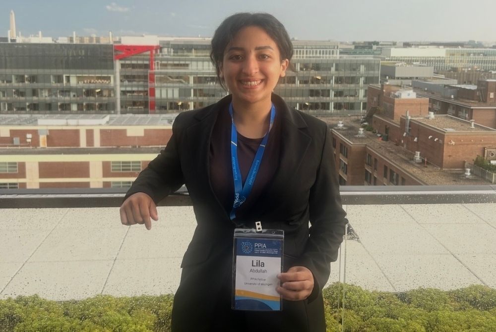 A JMC student, Lila Abdullah, identified by her name tag as a PPIA fellow from the University of Michigan, stands on a rooftop terrace smiling. She is wearing professional attire and holding up her event badge while posing with a cityscape backdrop featuring modern buildings.