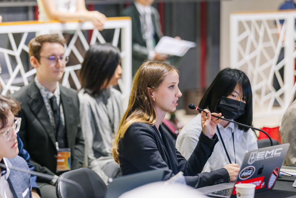 A JMC student with long hair is seated and actively speaking into a handheld microphone while looking forward. She wears a dark blazer and has a laptop in front of her, surrounded by other attendees in what appears to be a professional or academic event.