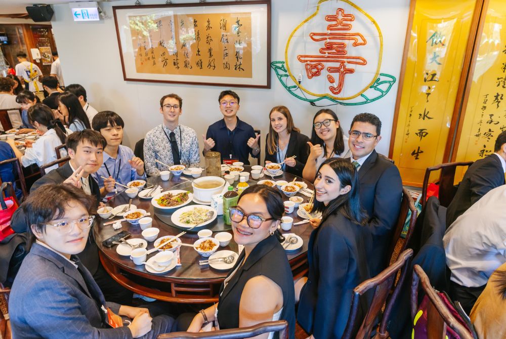 A group of professionals, dressed in business attire, are seated around a round dining table filled with various Chinese dishes, smiling and posing for the camera. The setting is an authentic traditional Chinese restaurant.
