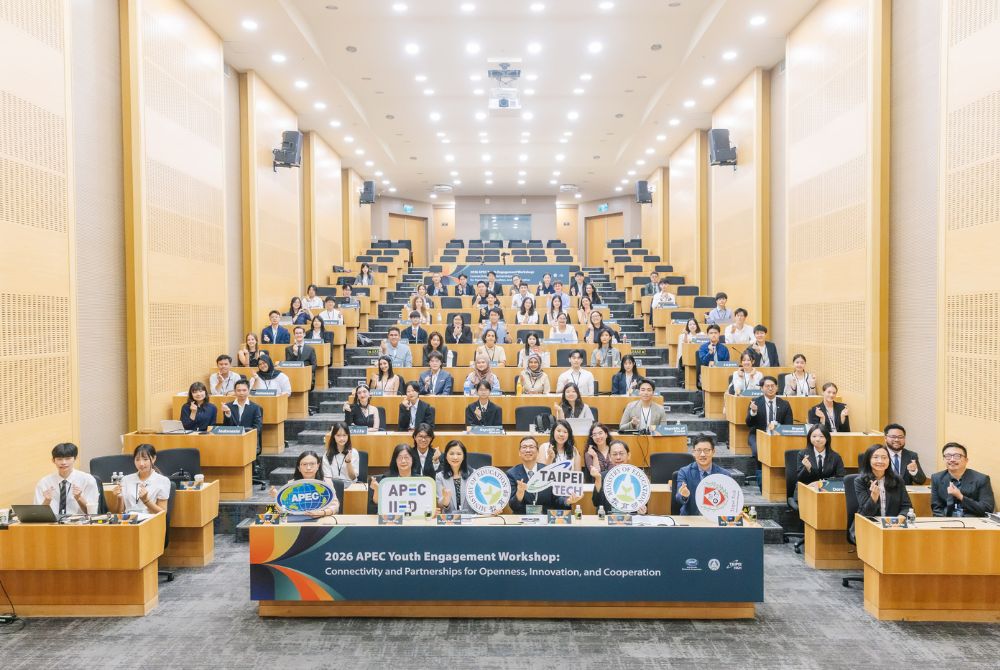 A group of young professionals and students are seated in a large, modern conference hall, participating in the 2026 APEC Youth Engagement Workshop. They are positioned at tiered desks, with some holding signs that represent their organizations or affiliations, while others gesture toward the camera.