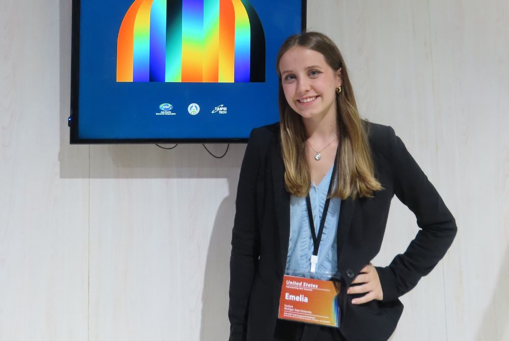 JMC student with a name tag stands confidently in front of a large screen displaying colorful graphics and logos. She is dressed professionally, wearing a black blazer over a light blue top, smiling warmly while posing for the photo.