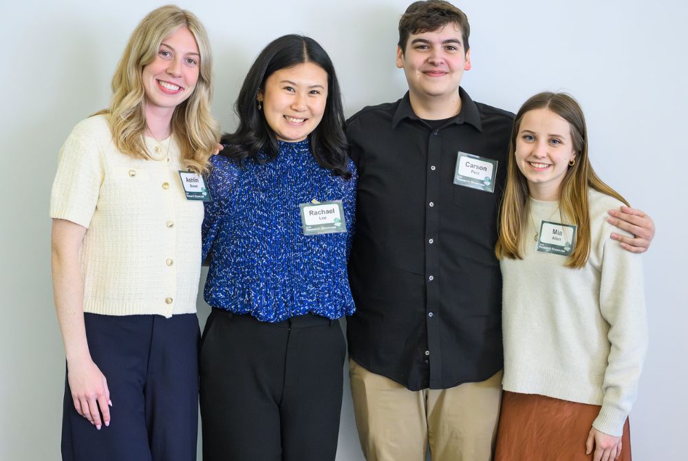 Four James Madison College students stand side by side, smiling at the camera, each wearing a name tag. They are positioned against a plain white wall, dressed in casual to smart-casual attire.