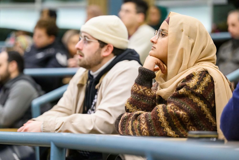 A young woman wearing a beige hijab and glasses sits attentively, her hand resting on her chin, while a man beside her, also wearing glasses, looks forward. They are seated among other students in what appears to be an academic lecture hall.