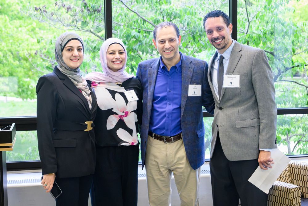Four individuals, two women wearing hijabs and two men, are standing together smiling for a photo. They appear to be attending a professional event, as suggested by their attire and name tags, with greenery visible through large windows behind them.
