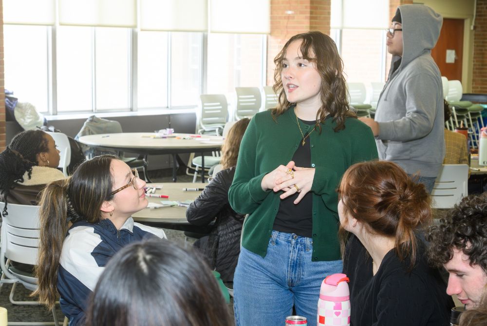 JMC senior Ava Parsons (standing, wearing a green sweater) talking with PRIME students, who are seated around a table in Case Hall.