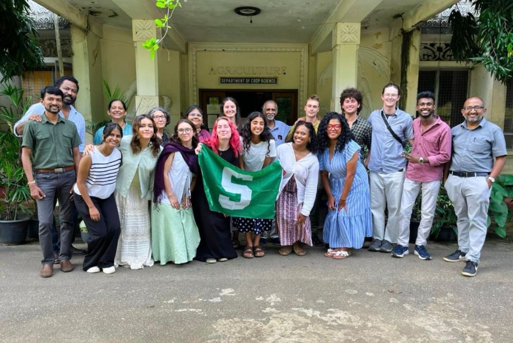 A group poses outside a university Department of Crop Science building, with several people holding a green flag marked with a large white “S.”