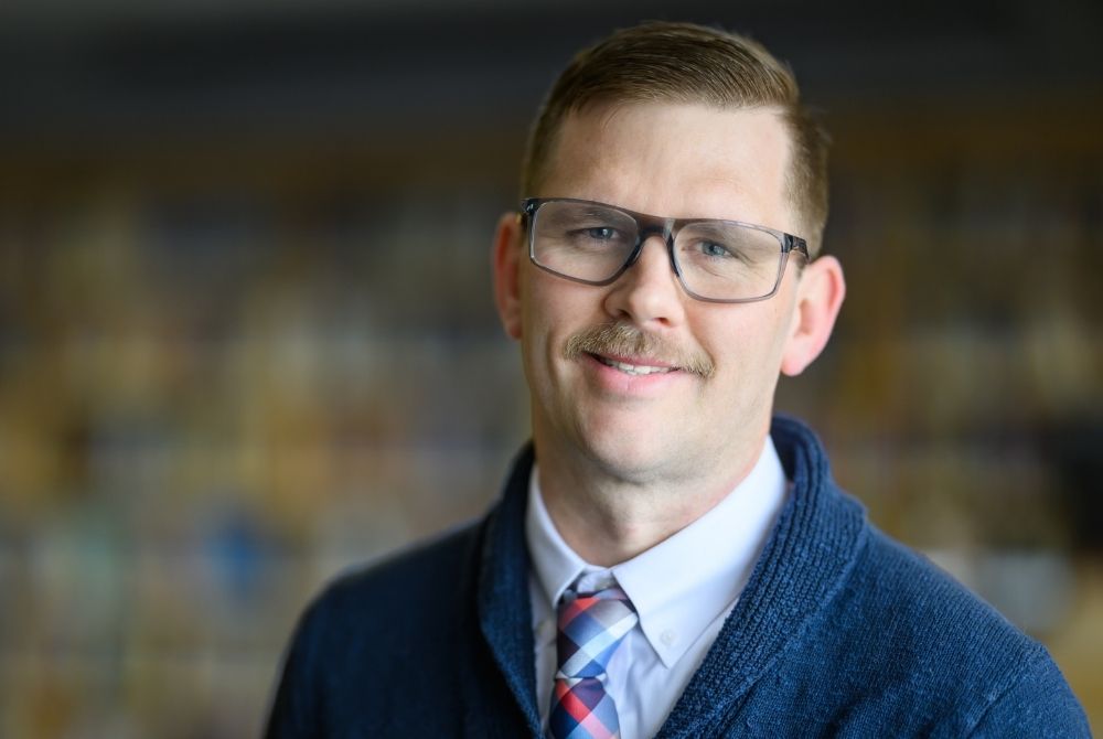 Sam Wright in the JMC Library, wearing a blue sweater, white collared shirt and plaid tie.