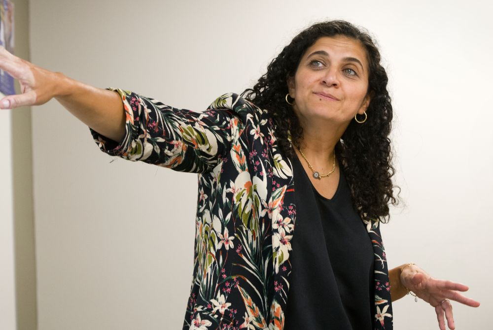 A woman with curly dark hair is standing indoors, extending her right arm outward while looking upward and smiling slightly. She wears a floral-patterned jacket over a black top, gold hoop earrings, and a necklace, with her left hand open in an expressive gesture.