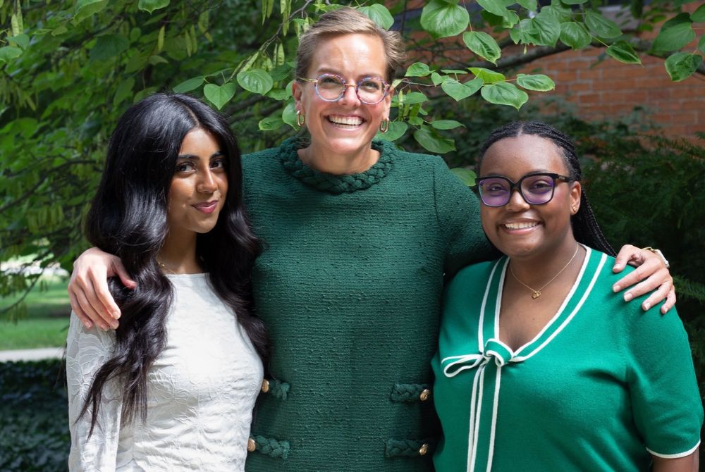Three people stand together outdoors, smiling warmly at the camera. The central woman has her arms around the other two, who are dressed in green and white outfits, with lush green foliage behind them.