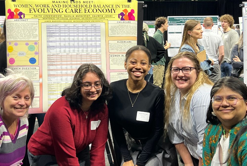 A group of three JMC students and two JMC professors pose together in front of a poster presentation inside of the Breslin Center. Behind them is a yellow presentation board presenting their work.