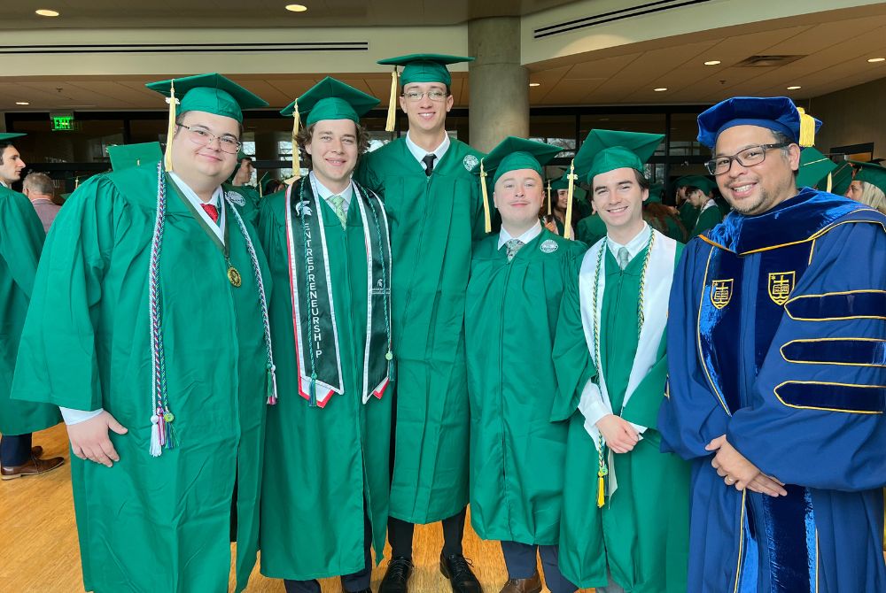 A group of five James Madison College graduates, dressed in green graduation gowns and caps, pose for a photo indoors. One JMC professor on the far right wears a blue academic robe with gold trim.