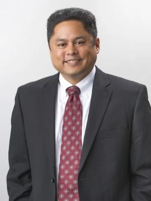 A JMC alumnus with short dark hair is smiling while wearing a black suit, white shirt, and patterned red tie. He stands against a plain light gray background, suggesting a professional portrait setting.