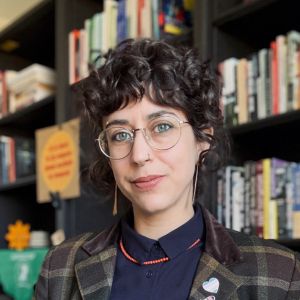 A JMC professor wearing round glasses and a plaid jacket stands in front of a book-filled shelf. Her dark curly hair frames her face as she looks directly at the camera.