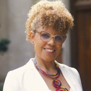An MSU professor with curly blonde hair and glasses smiles warmly at the camera. She wears a white blazer over a colorful beaded necklace, standing outdoors near a stone building.