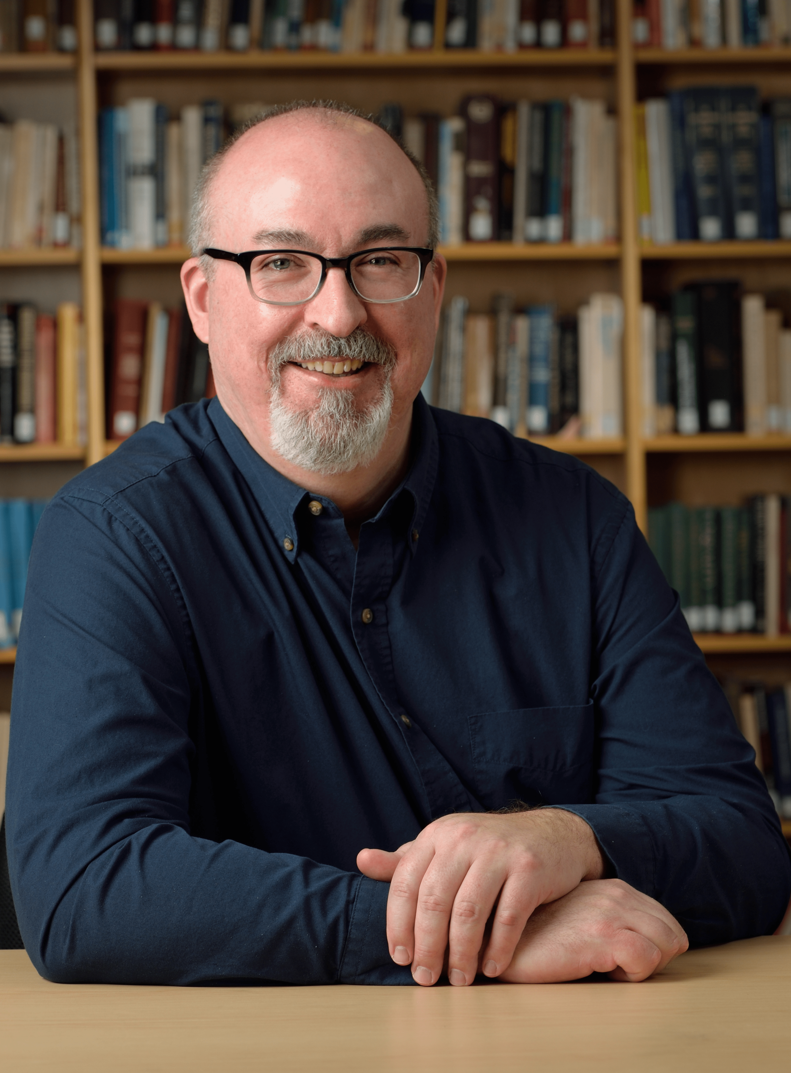 This photo shows Dean Cameron Thies in the James Madison College Library. He is wearing a blue collared shirt and his hands are folded in front of him.