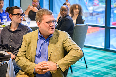 Students in the background and alumnus Troy Calkins listen to speakers at an Alumni Mixer