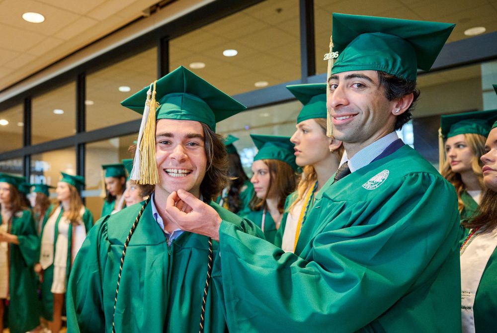 Two JMC graduates pose together in green caps and gowns, smiling at the camera.