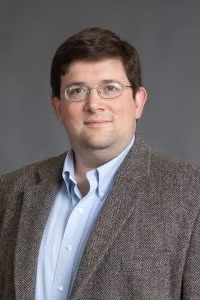 Headshot of Joshua Chafetz wearing glasses, a light blue shirt, and a brown blazer against a gray background.