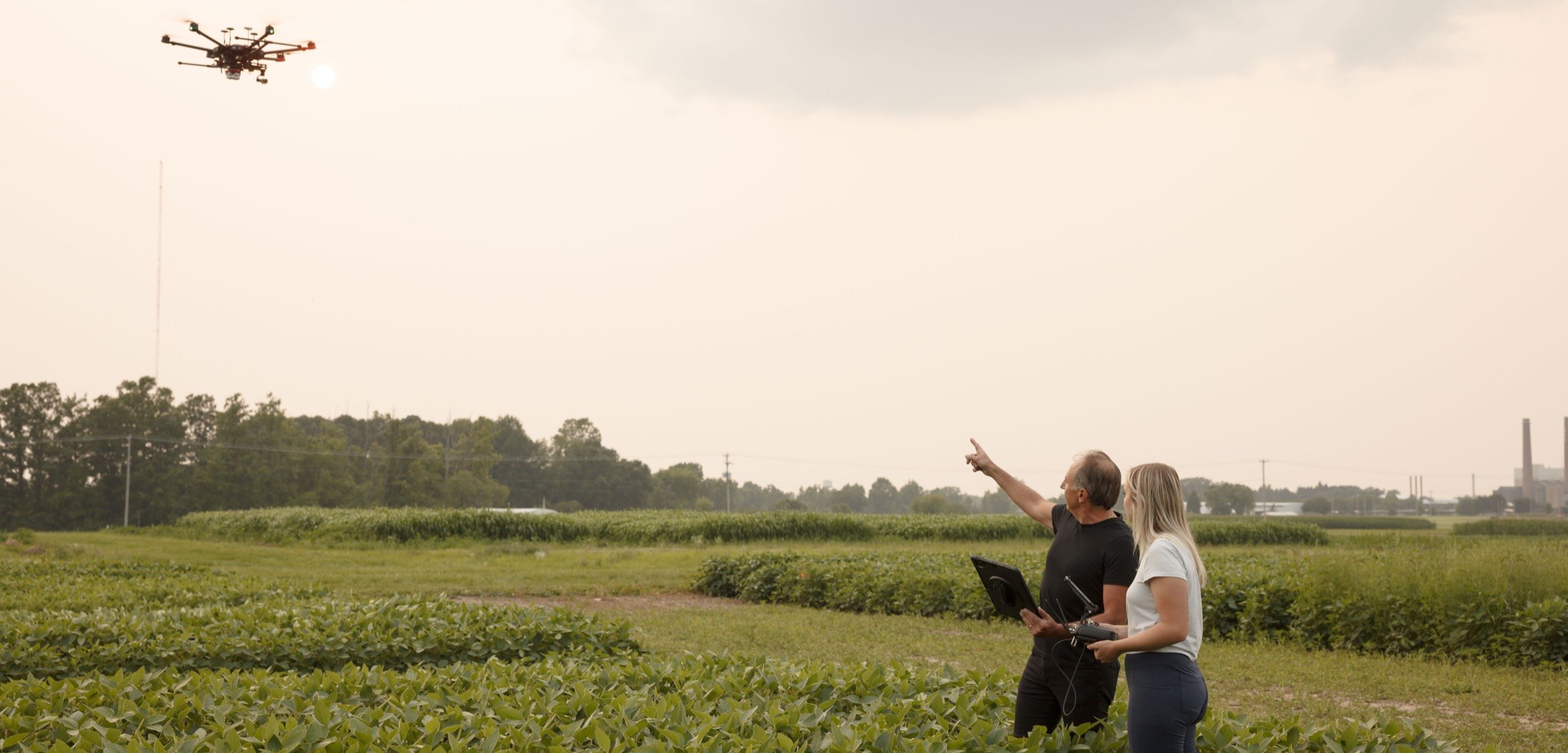 Two people stand in a field, one pointing at a drone in the sky. They appear to be monitoring it.