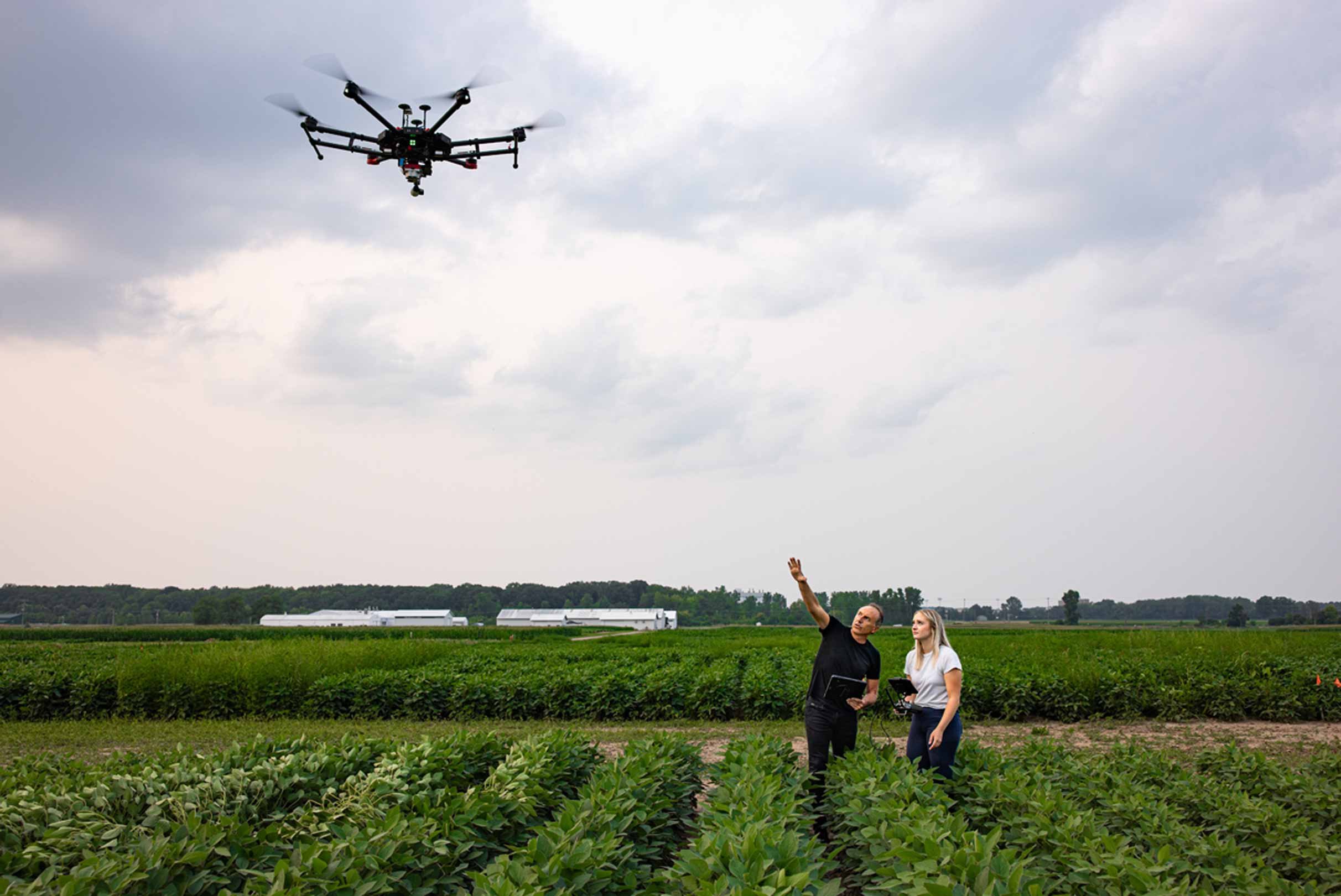 Two people in a field operating a drone with a tablet, with overcast skies above and industrial structures in the distance.