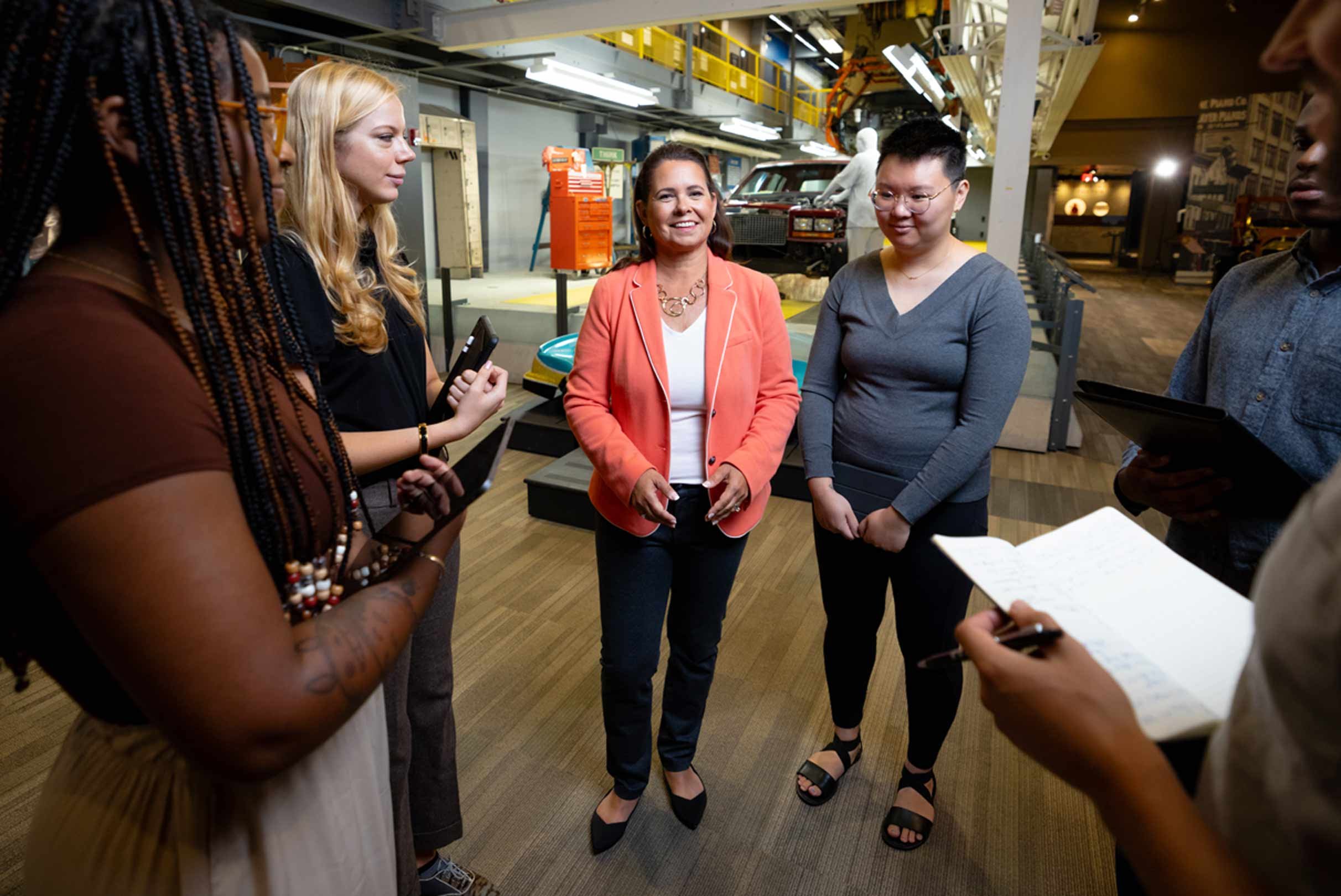 A group of individuals in a discussion with one person at the center speaking, in an industrial setting with machinery in the background.