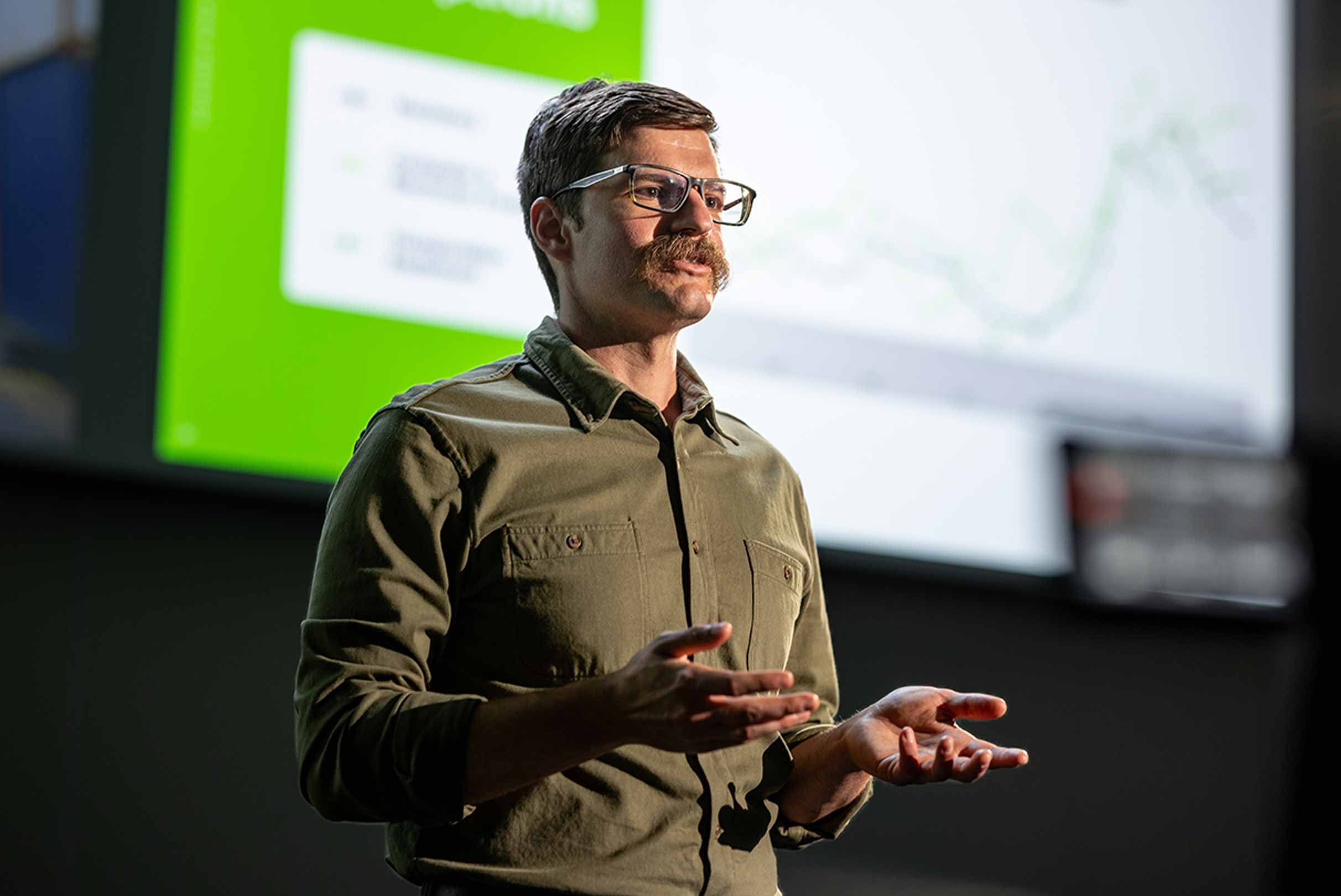  An individual presenting in front of a screen with a graph, gesturing with hands, in a lecture or meeting environment.